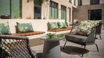 An outdoor seating area with wicker chairs and cushions, a central planter with greenery, and a building with large windows in the background.