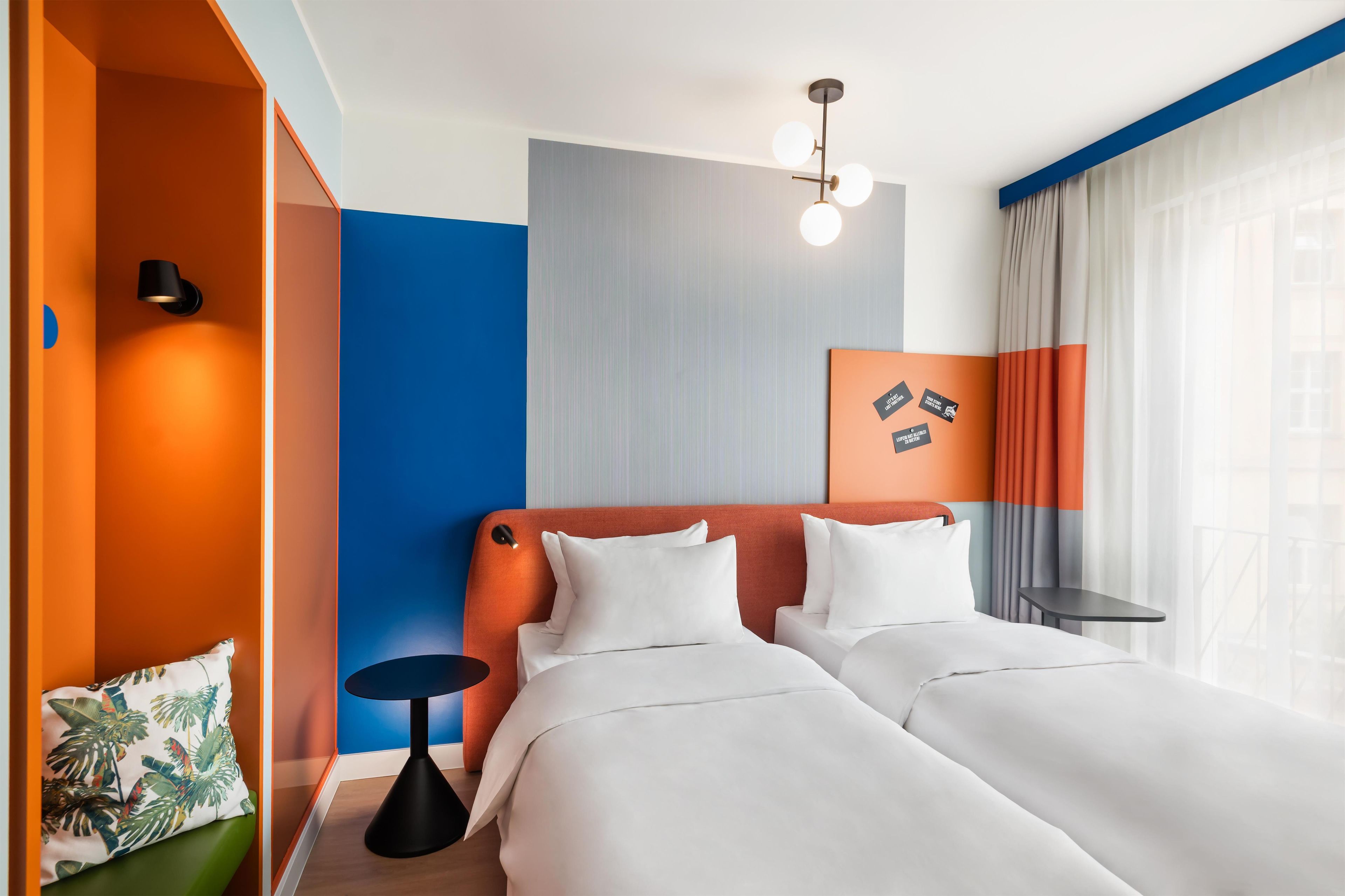 A hotel room with two beds, a small table, a wall-mounted light fixture, and a window with curtains.