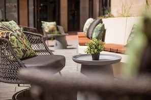 An outdoor seating area with wicker furniture, cushions, and a potted plant on a round table. - Me and All Hotel Leipzig, by Hyatt (Leipzig)