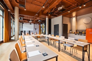 Sala de conferências com uma mesa grande, cadeiras, tela de projetor e um espaço de cozinha com exposição de frutas.