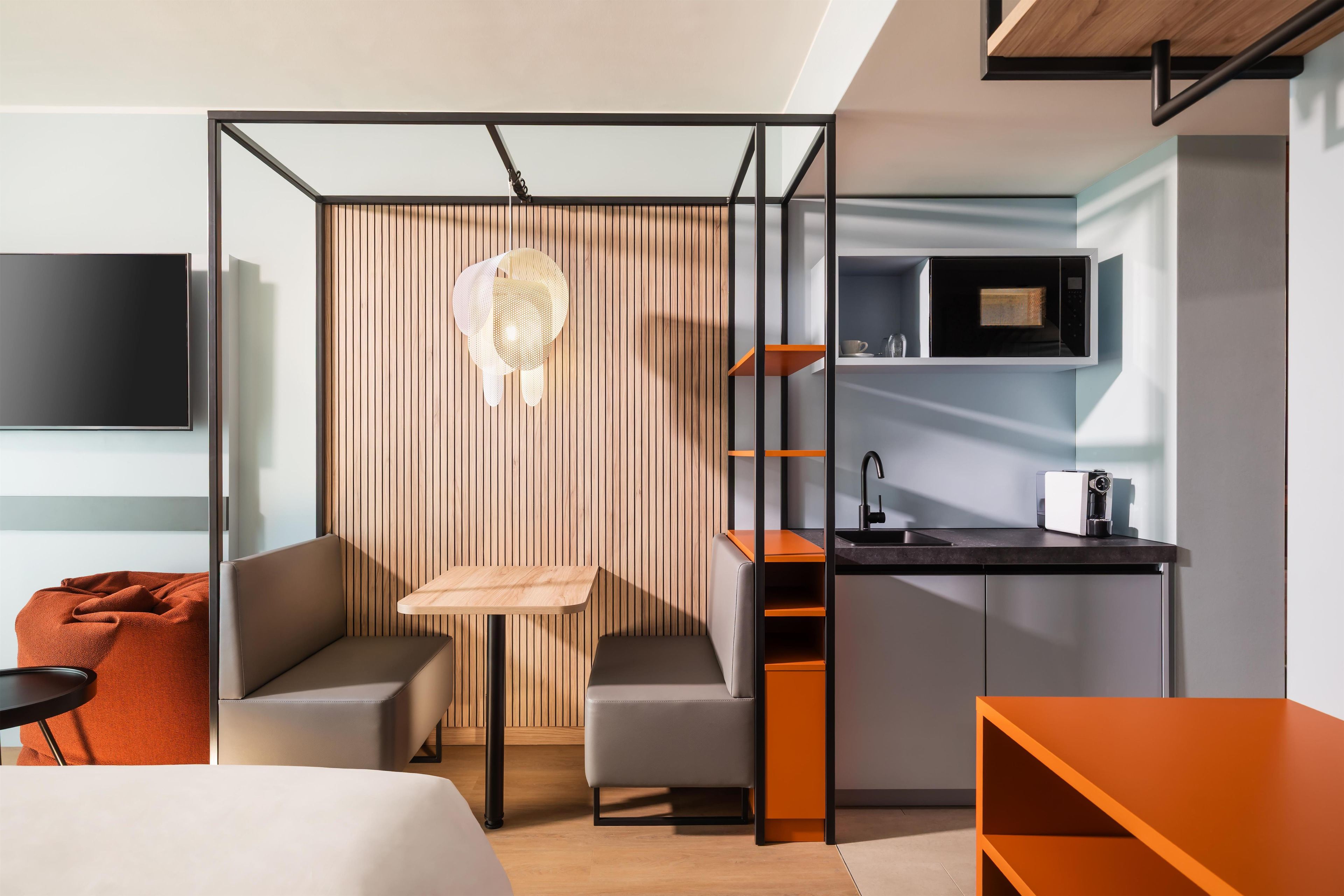 A modern hotel room with a kitchenette, a television, a seating area with a sofa and a small table, and a decorative hanging light fixture.