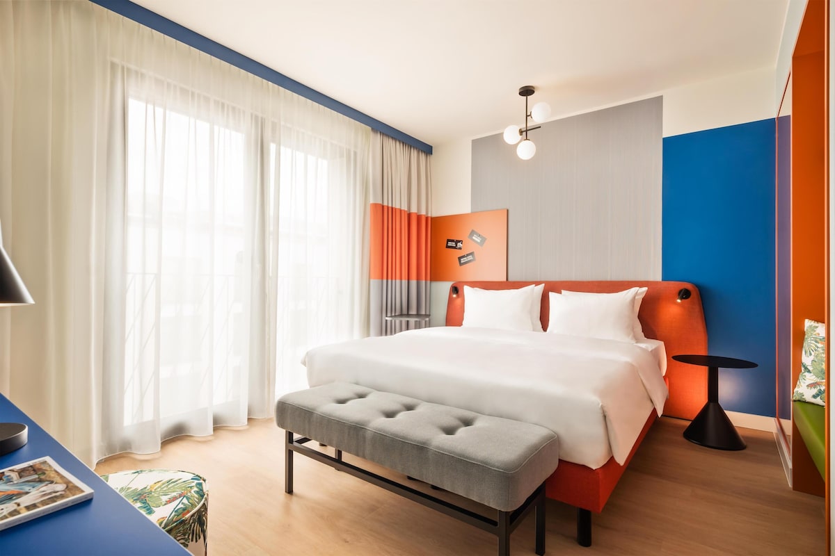 Habitación de hotel con cama, escritorio, silla, mesita y ventana con cortinas.