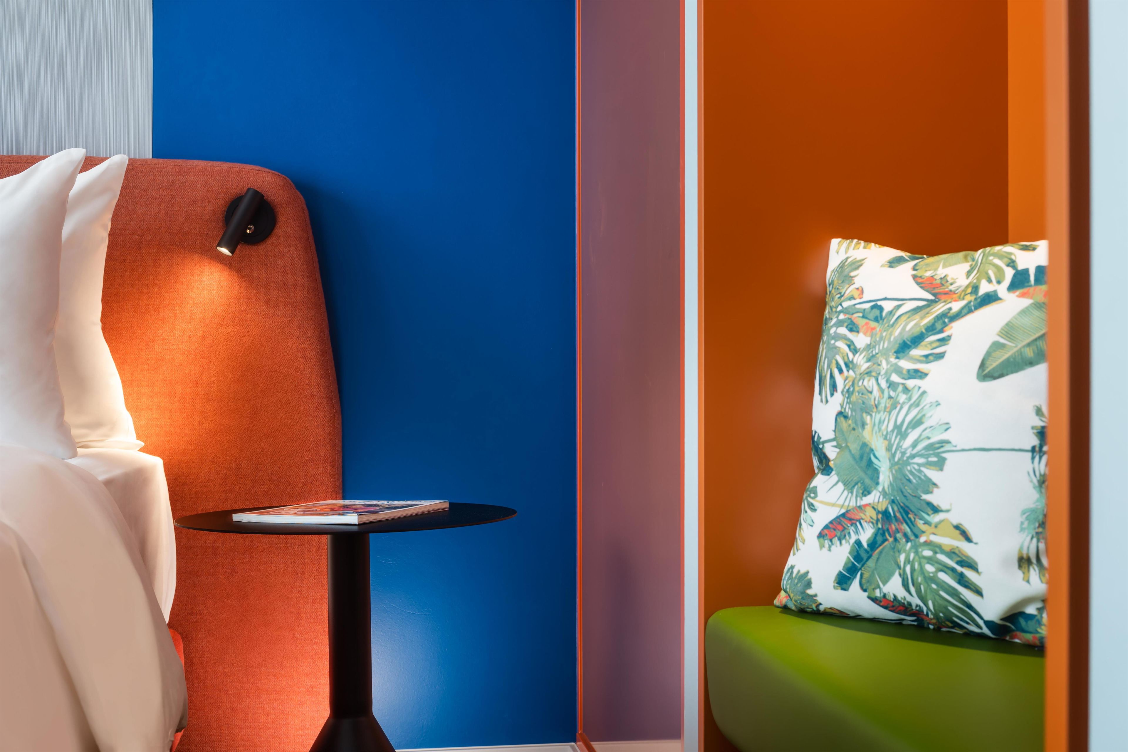 Una habitación con pared azul, una silla roja, una mesita redonda con una revista, y un sofá verde con un cojín estampado.