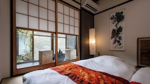 Een traditionele Japanse kamer met tatami vloerbedekking, een verschuifbare shoji-scherm, een bed met een deken met rood en gouden dessin, een wandairconditioner en een bloemwerk kunstwerk dat aan de muur hangt.