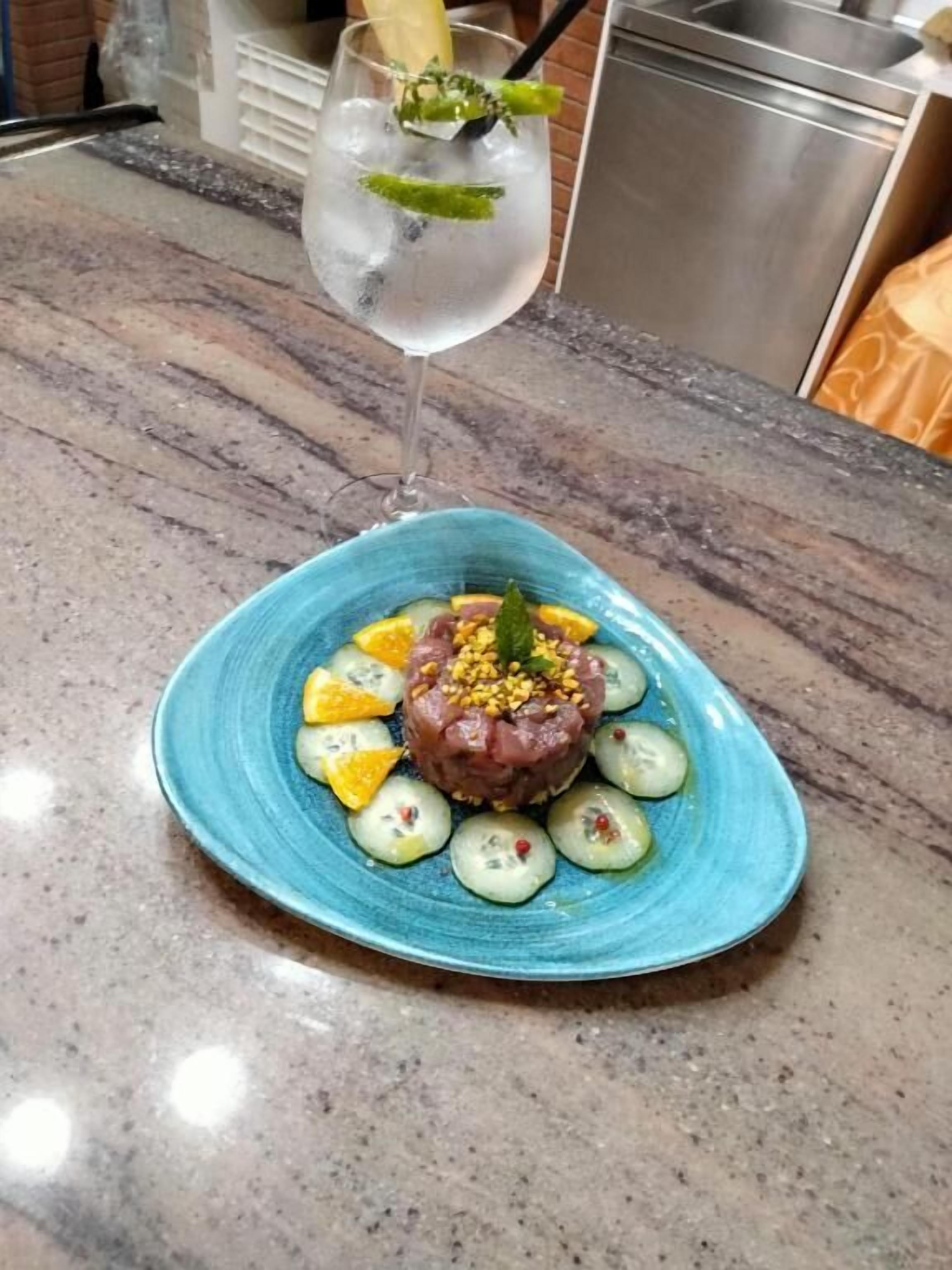 A blue plate with a dish of raw tuna tartare, cucumber slices, and yellow fruit, accompanied by a glass of white wine with a mint garnish.