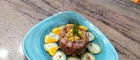 A blue plate with a dish of raw tuna tartare, cucumber slices, and yellow fruit, accompanied by a glass of white wine with a mint garnish.