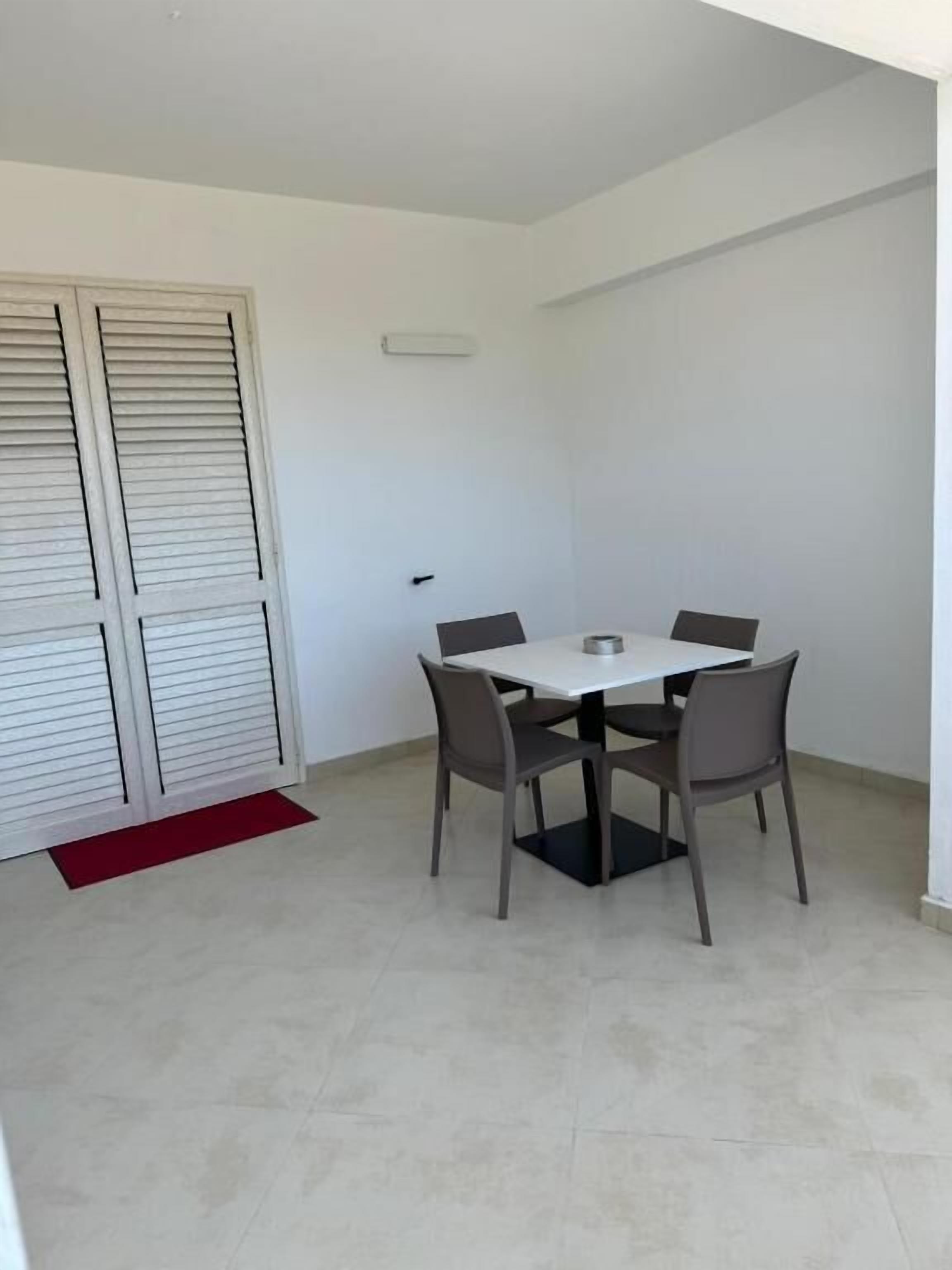 Une petite pièce propre, avec une table blanche et quatre chaises, un tapis rouge et une porte blanche avec des volets.