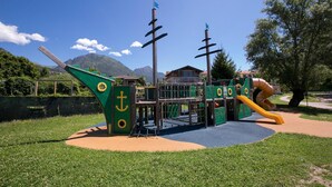 Un parque infantil con un tobogán y estructuras para escalar, rodeado de césped y árboles, con montañas al fondo.
