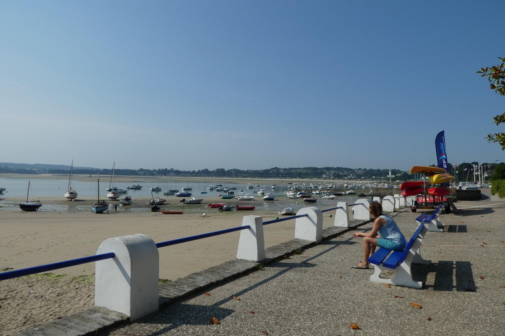Une plage avec des bateaux, une personne assise sur un banc et un drapeau.