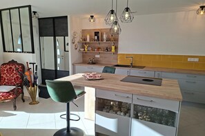 A modern kitchen with a central island, pendant lights, and a dining area with a red upholstered chair. - Ground floor holiday home in Plestin des Grèves (Plestin les Grèves)
