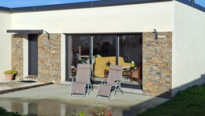 Un bâtiment moderne agrémenté d’éléments en pierre, une terrasse avec deux chaises et un intérieur visible à travers de grandes portes vitrées.