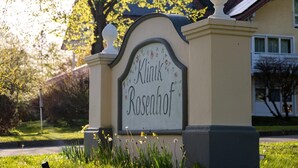 Ein Schild mit der Aufschrift 'Minik Rosenhof' vor Bäumen und einem Gebäude.