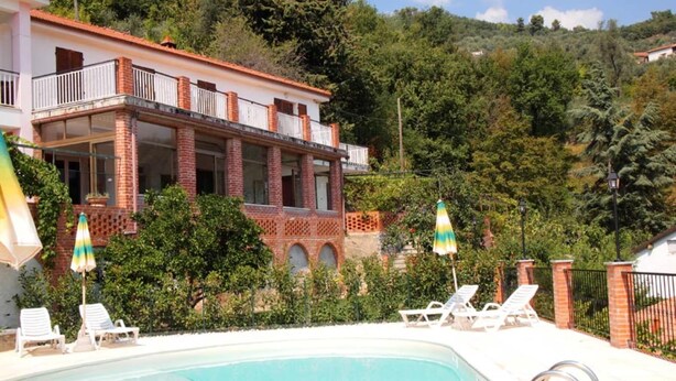 Ein Poolbereich mit Liegestühlen und Sonnenschirmen, ein Gebäude mit Balkon sowie eine hügelige Landschaft im Hintergrund.