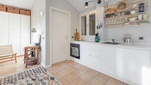 A modern kitchen with white cabinetry, a built-in oven, and a countertop with a kettle and a coffee maker. There is a wicker basket on the floor and a chair in the corner. - Studio in Porlezza am Luganersee (Porlezza)