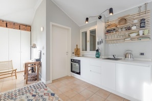 A modern kitchen with white cabinetry, a built-in oven, and a countertop with a kettle and a coffee maker. There is a wicker basket on the floor and a chair in the corner. - Studio in Porlezza am Luganersee (Porlezza)