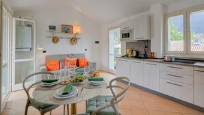 A modern kitchen with white cabinets, a dining area with a table set for two, and a view of the outdoors through large windows. - Studio in Porlezza am Luganersee (Porlezza)