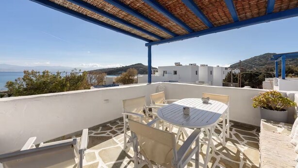 A terrace with a table and chairs, a potted plant, and a view of the sea and buildings. - Paros 3 bedrooms Maisonette for 6 persons by MPS (Molos)