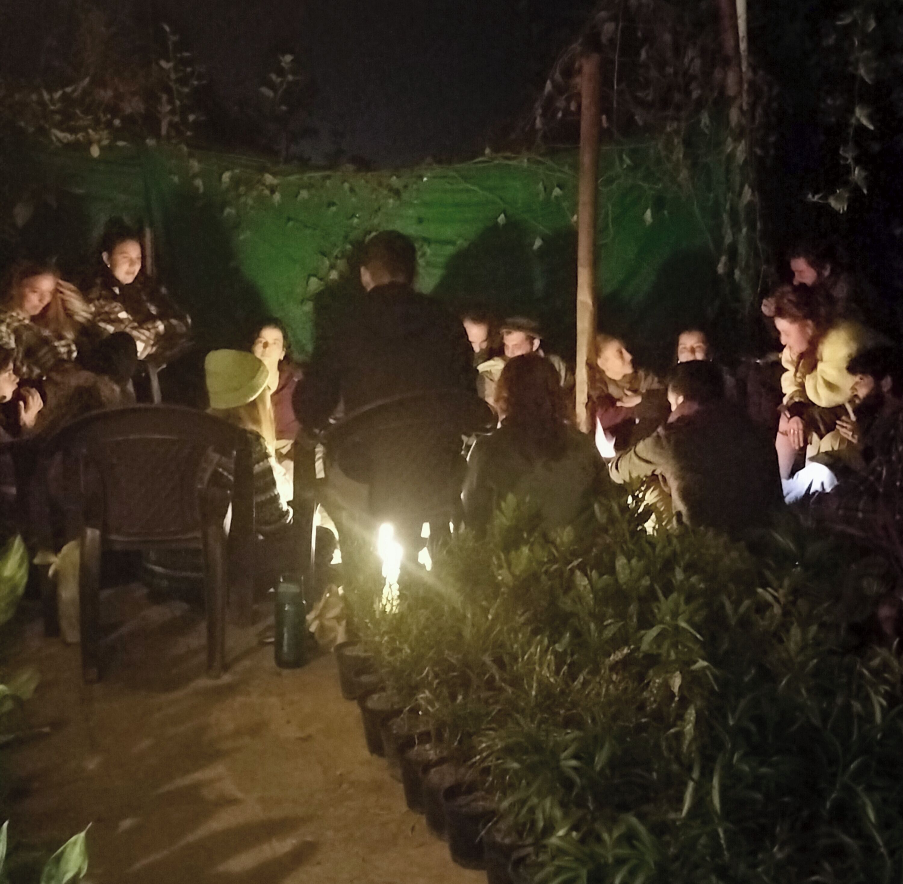 Eine Gruppe von Menschen ist abends draußen versammelt und sitzt auf Stühlen und Bänken, im Hintergrund ist eine grüne Plane zu sehen.