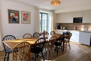 Un coin repas avec une table et des chaises en bois, une cuisine avec des meubles blancs et des appareils électroménagers, et deux œuvres d’art florales encadrées accrochées au mur.