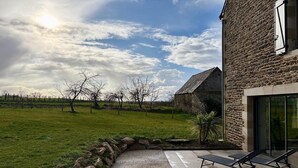 A stone building with a modern extension, a gravel patio with lounge chairs, and a grassy field with trees in the background. - Lovely holiday cottage in Corseul (Corseul)