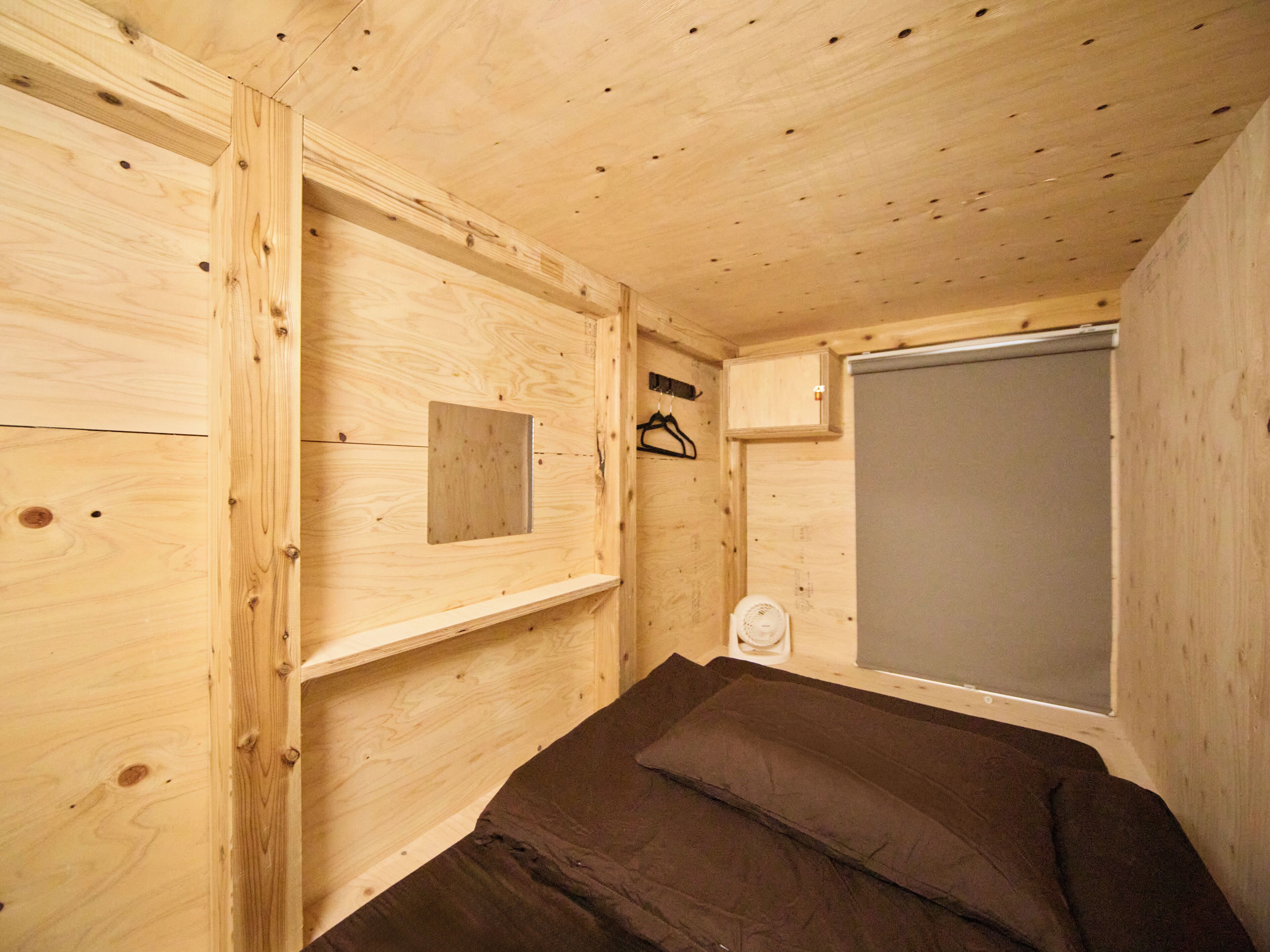 A compact wooden room with a bed, a small shelf, a cabinet, and a window.