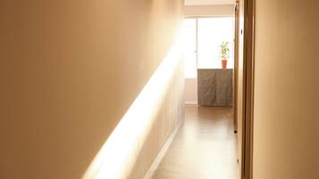 A narrow hallway with a wooden floor and light-colored walls. A window at the end of the hallway allows natural light to enter.