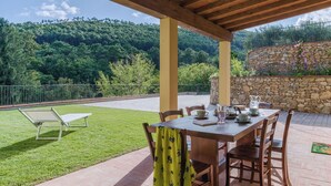 Alpendre coberto com mesa e cadeiras, gramado e muro de pedra com vista para uma encosta arborizada.