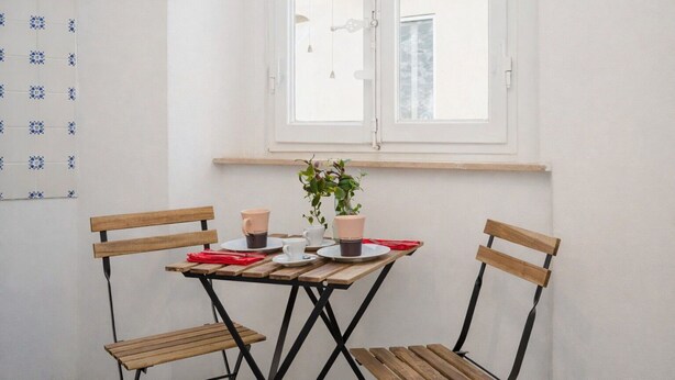Ein kleiner, privater Sitzbereich im Freien mit Tisch und zwei Holzstühlen, einem Blumentopf und einem Fenster mit Blick auf die Straße.