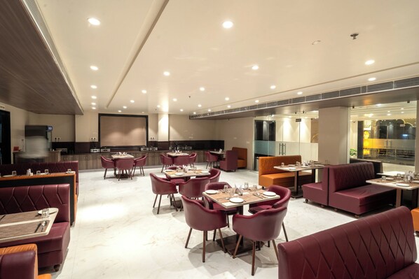 A modern dining area with multiple seating options including booths and tables, a kitchen area in the background, and a well-lit ceiling with recessed lighting.
