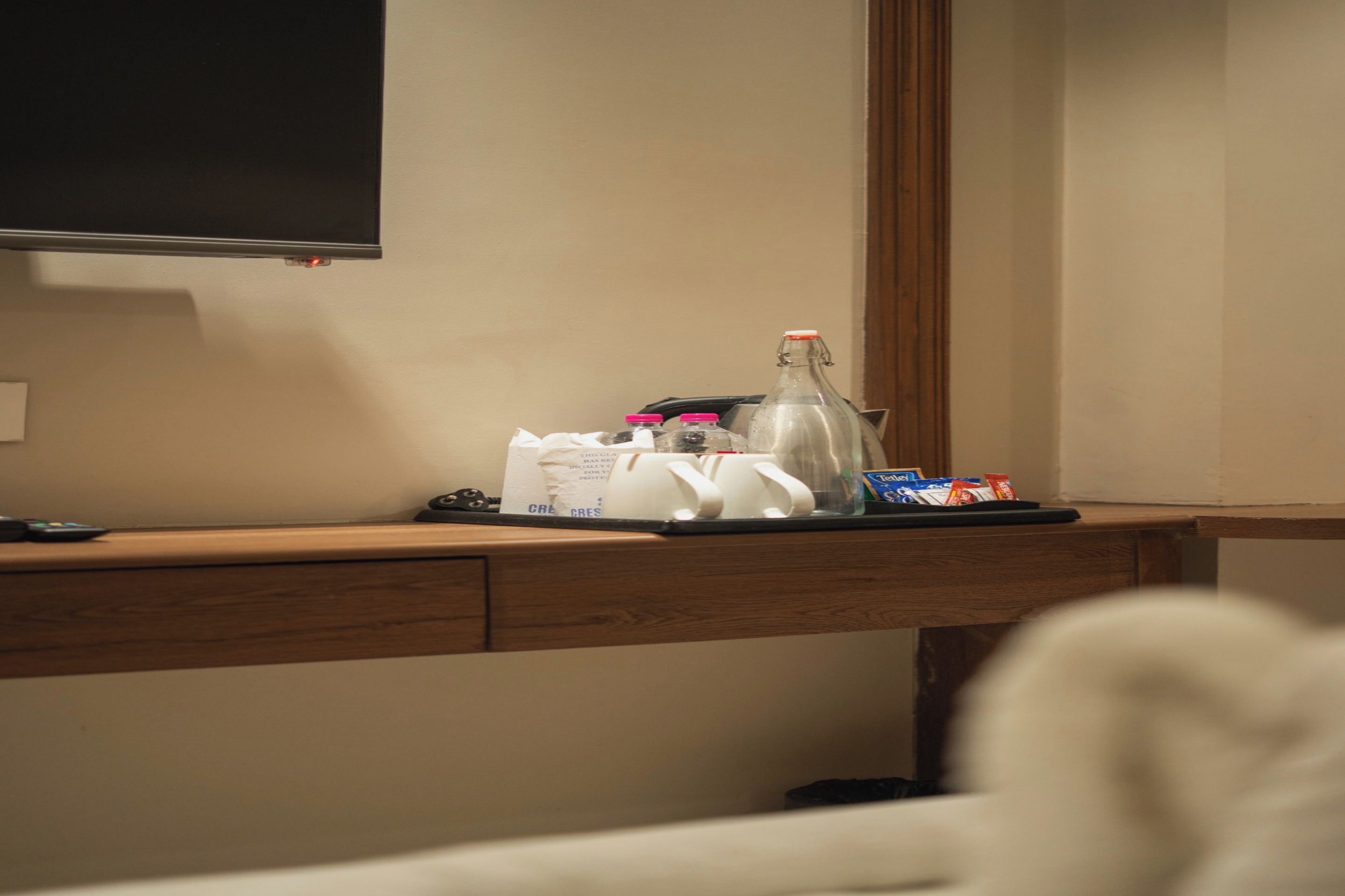 Mostrador de hotel con televisor de pantalla plana en la pared, una bandeja con artículos de aseo, una botella y algunos aperitivos.