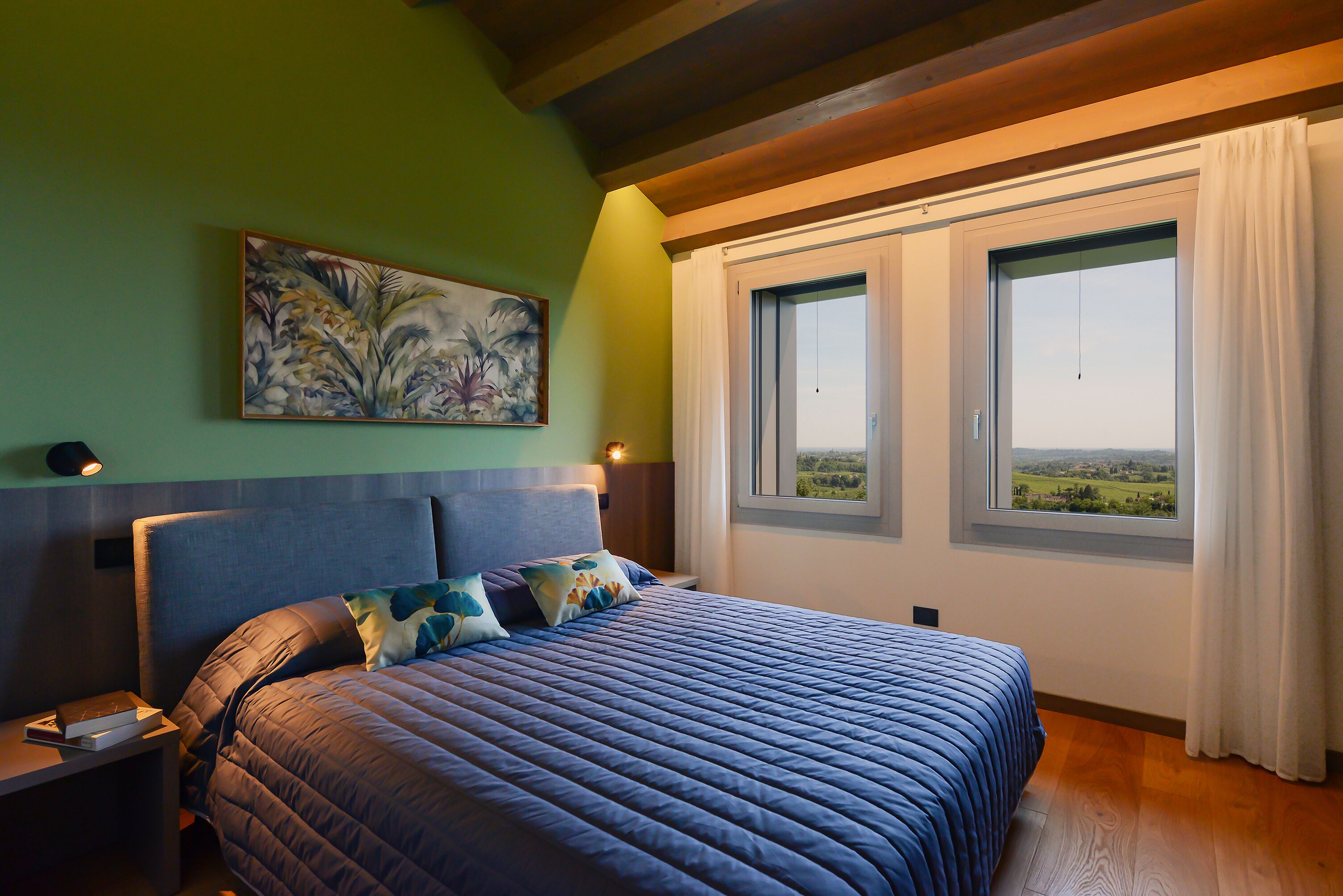 Een slaapkamer met een groot bed, een nachtkastje, een schilderij aan de muur en een raam met uitzicht op het landschap.