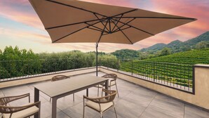 Una terraza con una mesa y sillas, un gran paraguas y una vista panorámica de colinas verdes.