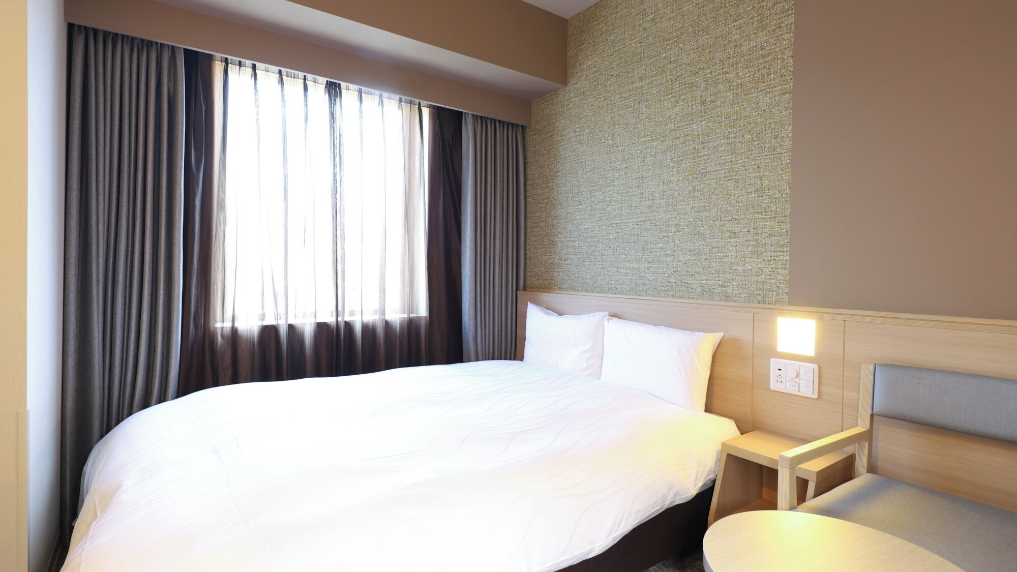 A hotel room with a large bed, a bedside table, a small round table, and a wall-mounted light.