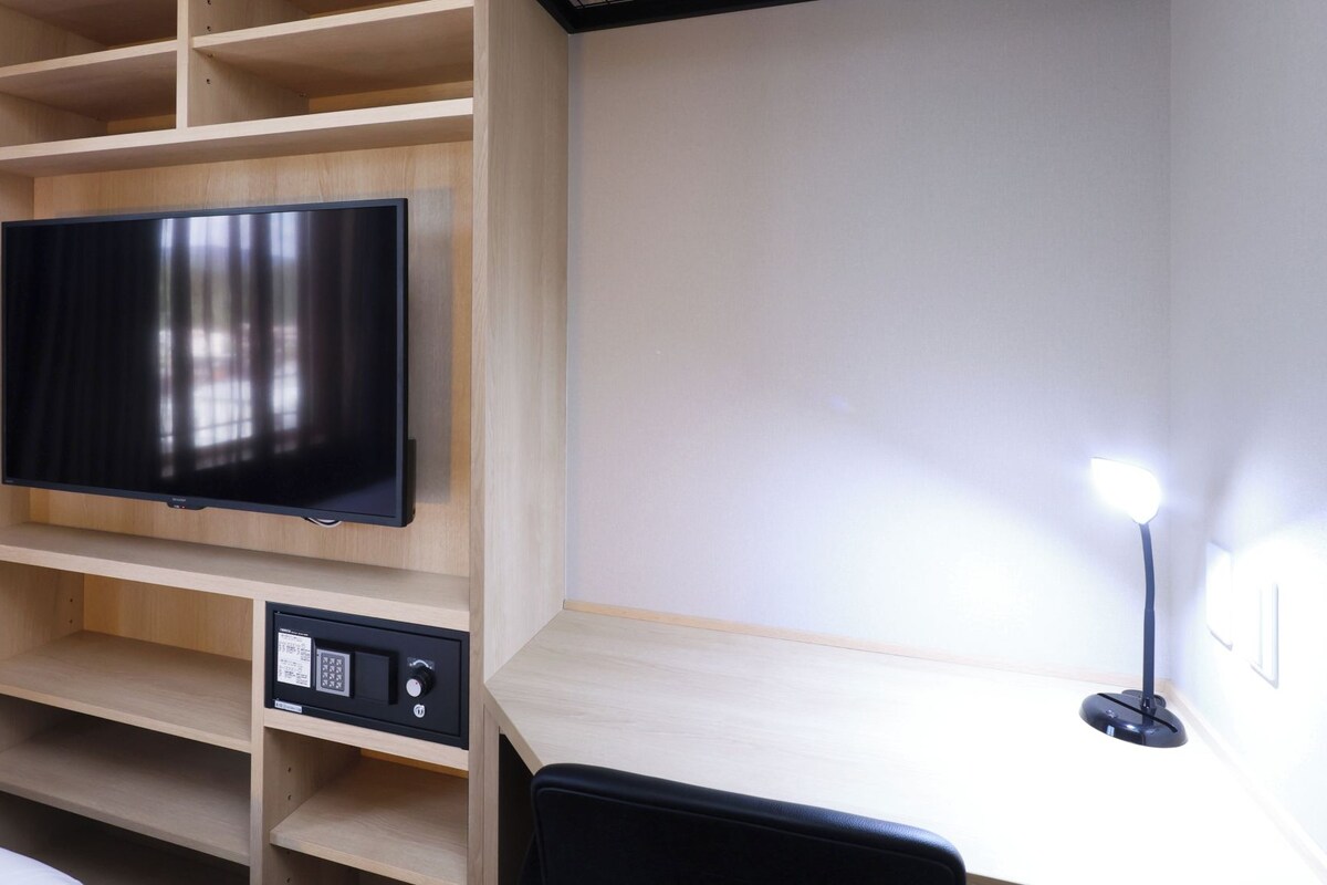 A room with a television mounted on a wooden shelf, a desk with a chair, and a wall-mounted light.