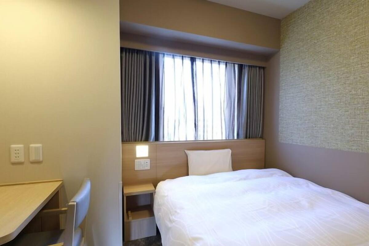 Habitación de hotel con cama, escritorio, mesita de noche, ventana con cortinas y pared con textura.