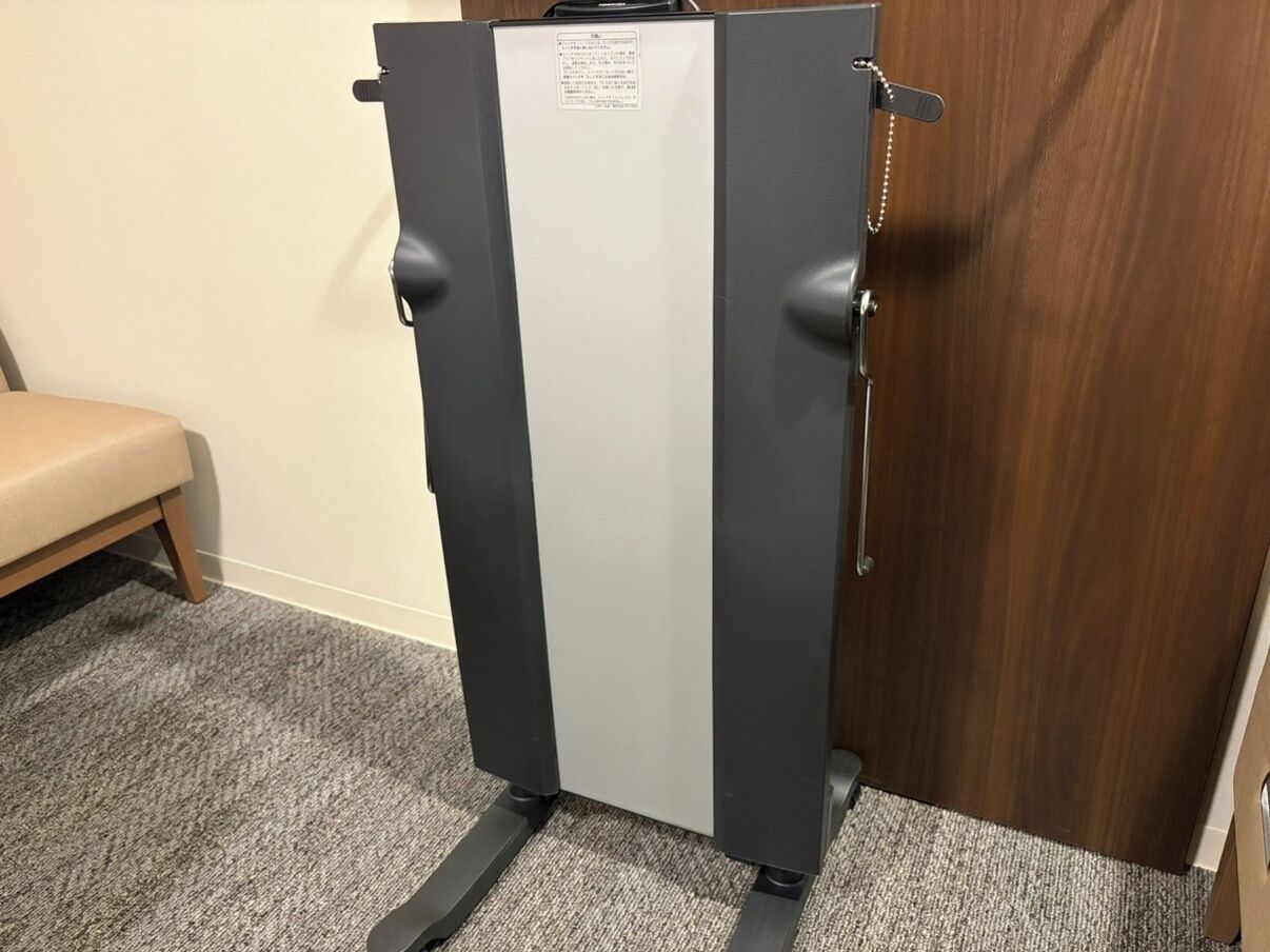 A compact, dark-colored refrigerator with a white top panel, positioned against a wooden door and a beige bench in a room with carpeted flooring.