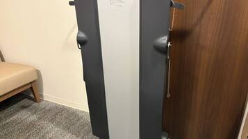 A compact, dark-colored refrigerator with a white top panel, positioned against a wooden door and a beige bench in a room with carpeted flooring.