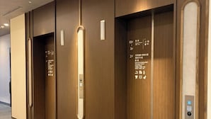 A hotel corridor with wooden paneling, a tiled floor, and three elevators with floor indicators. - Dormy Inn Tsuruga (TSURUGA SHI)