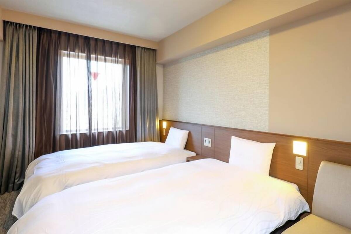 Habitación de hotel con dos camas, un ventanal con cortinas y una lámpara fijada a la pared.