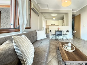 Ein modernes Wohnzimmer mit grauem Sofa, weißen, strukturierten Kissen, einem Holztisch und einem Essbereich mit kleinem Tisch und Stühlen.