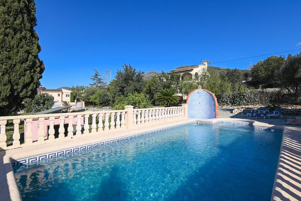 A clear blue swimming pool with a decorative fountain, surrounded by a white balustrade and lush greenery.