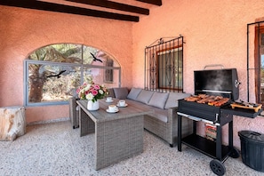 An outdoor patio with a wicker table, a sofa, a grill, and a window with a view of palm trees. - Finca La Solana – Holiday home with a private pool (Jalón)