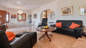 Ein geräumiges Wohnzimmer mit einer schwarzen Ledersofa, einem runden Holztisch und einer schwarzen Couch mit orangefarbenen Kissen. Es gibt eine Essecke mit einem weißen Tisch und Stühlen, sowie ein Bücherregal voller Bücher.