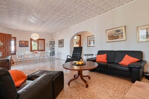 A spacious living area with a black leather sofa, a round wooden coffee table, and a black leather couch with orange cushions. There is a dining area with white chairs and a table, and a bookshelf filled with books. The room has a high ceiling with a decorative pattern and framed artwork on the walls. - Finca La Solana – Holiday home with a private pool (Jalón)