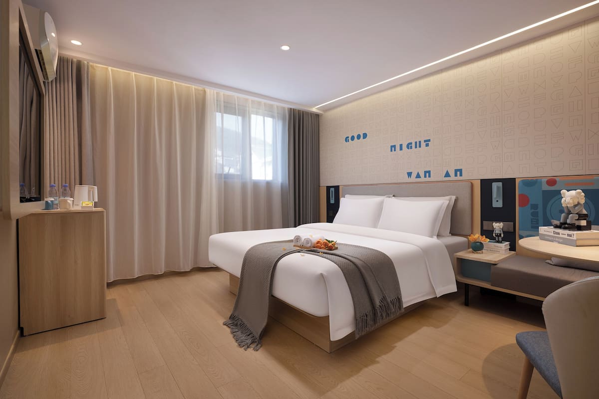A modern hotel room with a neatly made bed, a bedside table with a lamp, and a small desk with a chair.