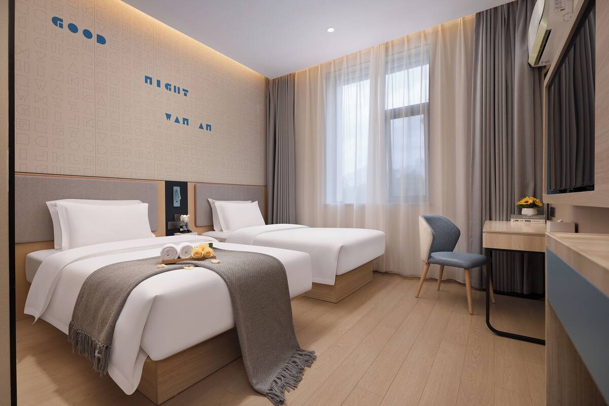 A hotel room with two beds, a desk, a chair, and a large window with curtains.