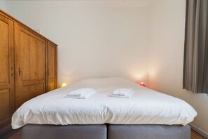 Ein ordentlich bezogenes Bett mit weißen Leinen und zwei gefalteten Handtüchern, ein Holzschrank und eine Nachttischlampe.