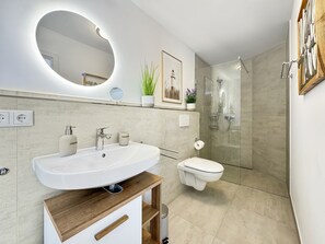 A modern bathroom with a large oval mirror, a sink, a toilet, and a glass-enclosed shower. - 5220 - Küstenfeuer - Coastal Lighthouse - 5220 - Luxury - 3 bedrooms - in Dahme with ocean views (Dahme)