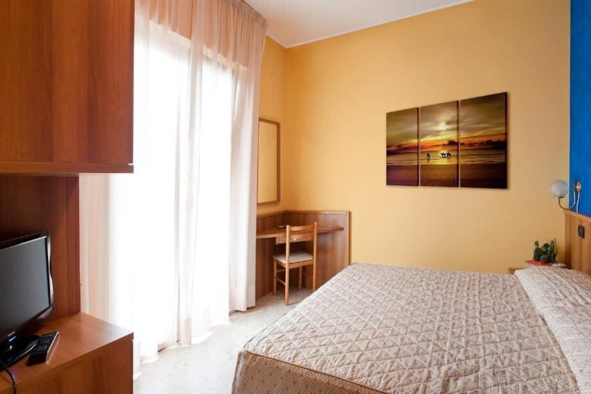 A hotel room with a bed, a desk, a television, and a painting on the wall.
