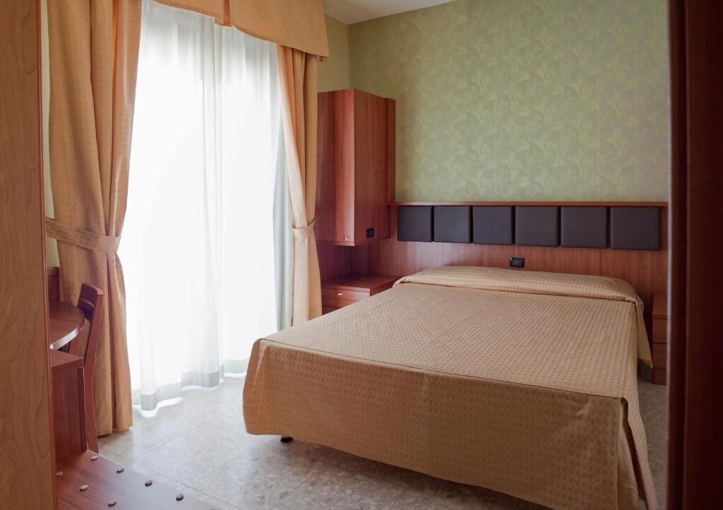 Une chambre d’hôtel avec un lit, une table de chevet en bois, une armoire et une fenêtre avec des rideaux.